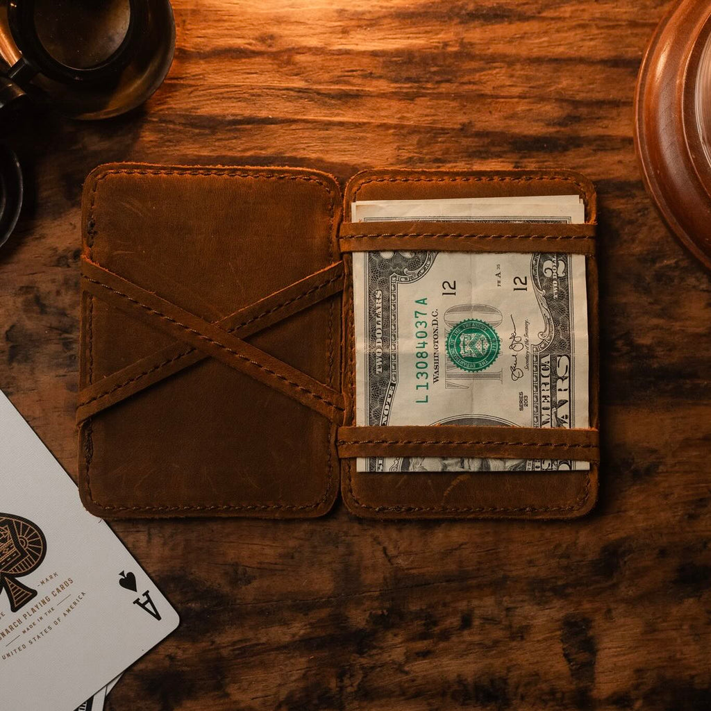Front Pocket Magic Wallet opened on a wooden table holding folded cash, beside playing cards and a glass