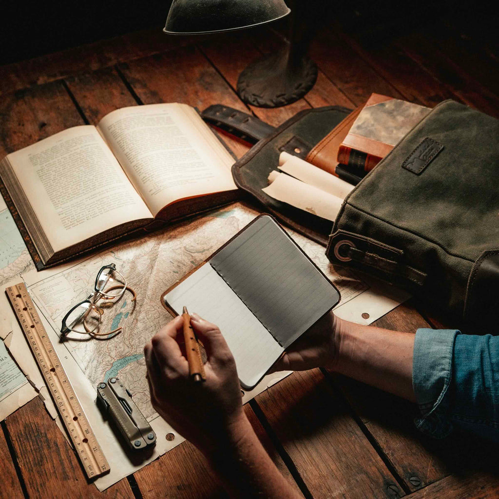 Leather Pocket Notebook in Black being written in at a desk with maps, books, glasses and a canvas bag