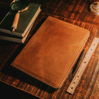 Old Design Medium Leather Portfolio in Tobacco on a wooden desk with books, a magnifying glass, pen and ruler