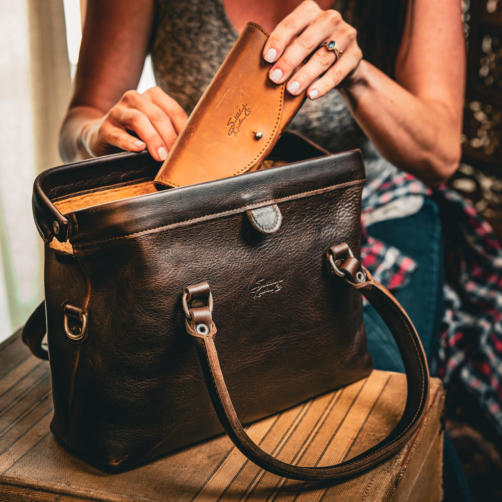 Everyday Purse in Dark Coffee Brown on a wooden stool as a woman tucks a tan leather wallet inside