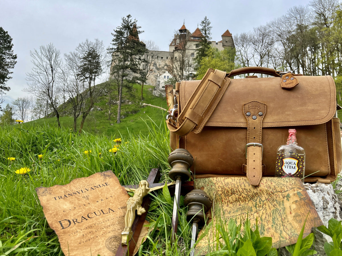 Tan leather slim briefcase in the grass below a hilltop castle, beside a Dracula book, stakes, map and holy water