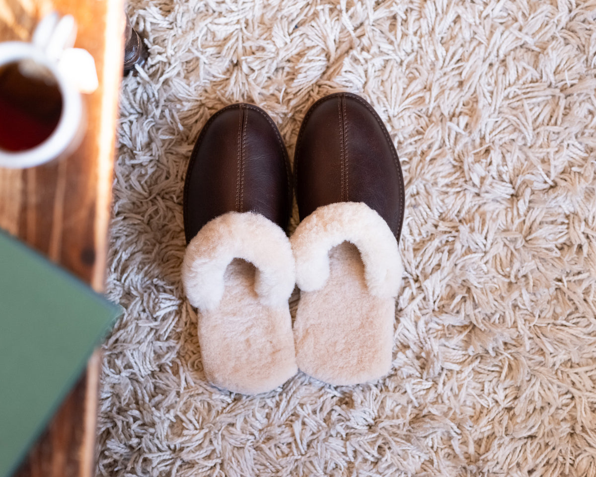 Sheepskin Slippers - Men - New Design in Dark Coffee Brown, top view of a pair on a shaggy rug beside a cup of tea
