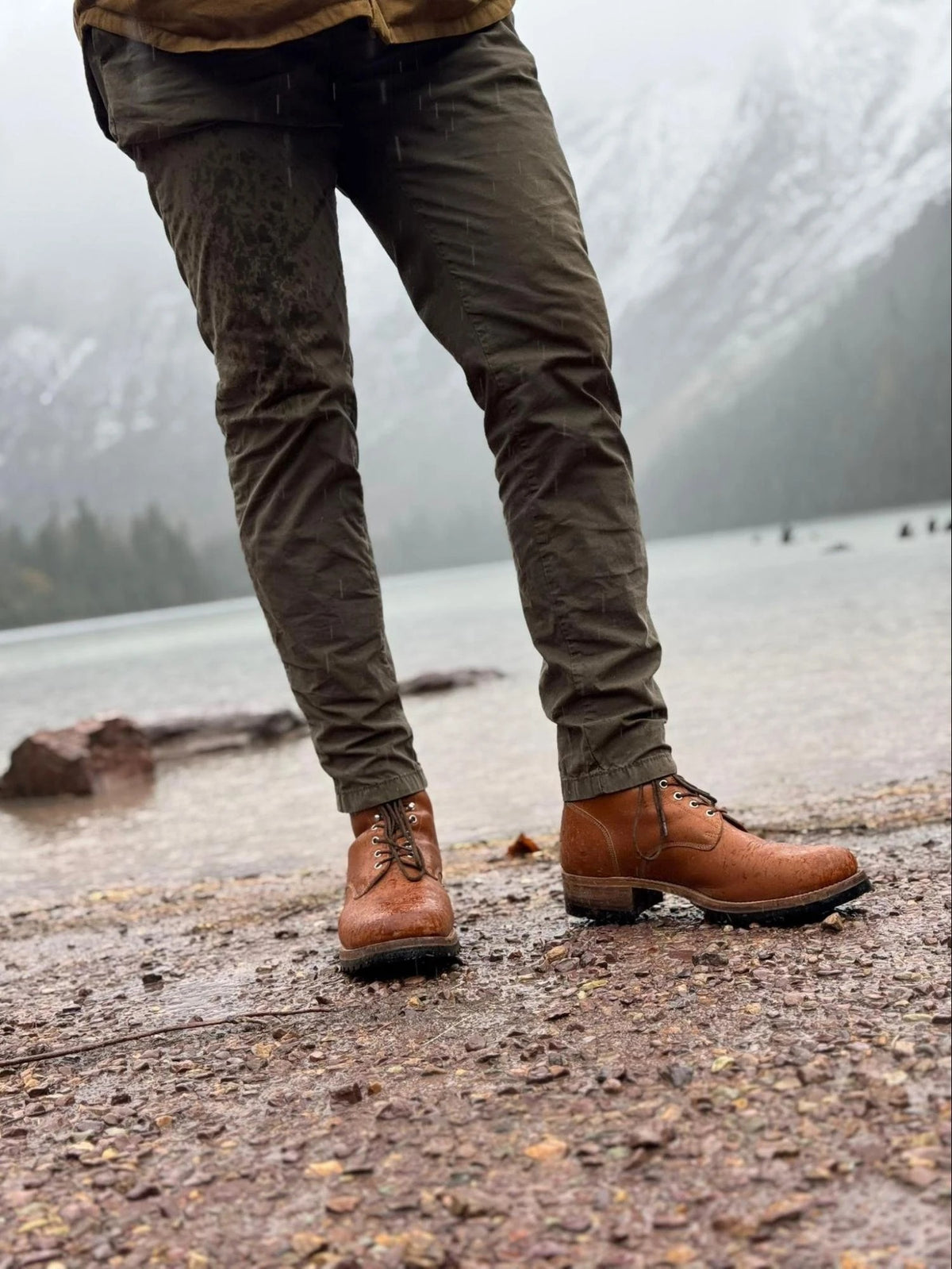 Huntsman Boots - Natural on a person in olive pants standing on a wet rocky lakeshore below misty mountains