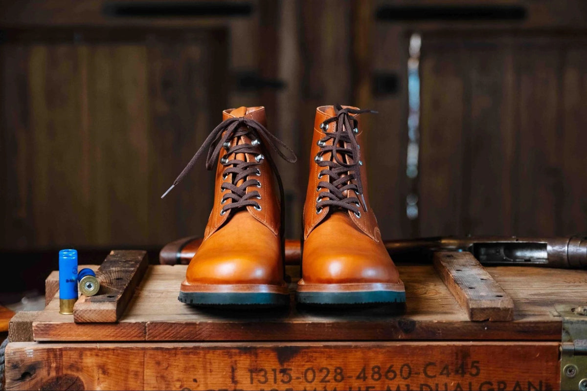 Huntsman Boots - Natural, front view of a laced pair on a stenciled wooden crate in a rustic cabin