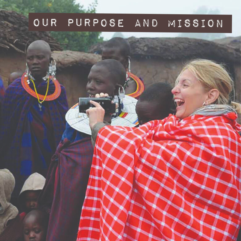 Our Purpose and Mission: a laughing woman wrapped in a red checked blanket films with a video camera beside Maasai women in beaded collars, outside mud houses.