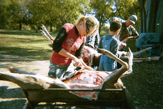Suzette and two children butchering a deer over a wheelbarrow in a park.