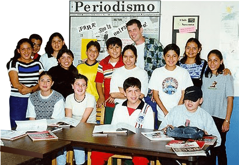 A class of Mexican schoolchildren posed around a table at Centro NOE, in front of a bulletin board headed Periodismo.