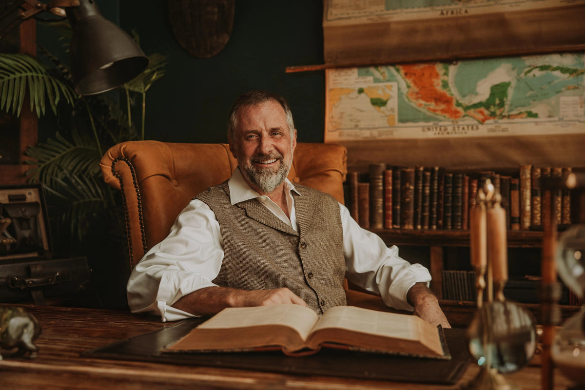 Smiling bearded man in a vest seated in a leather armchair at a desk with an open book, bookshelves and wall maps behind