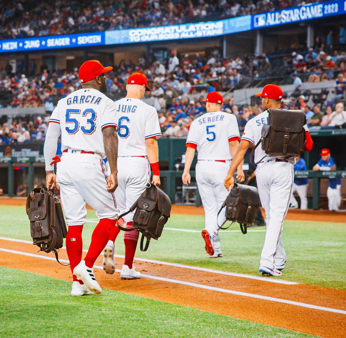 Garcia, Jung, Seager and Semien walk along the baseline in their Rangers uniforms, each carrying a Tank Leather Backpack, under a board congratulating the 2023 All-Stars.