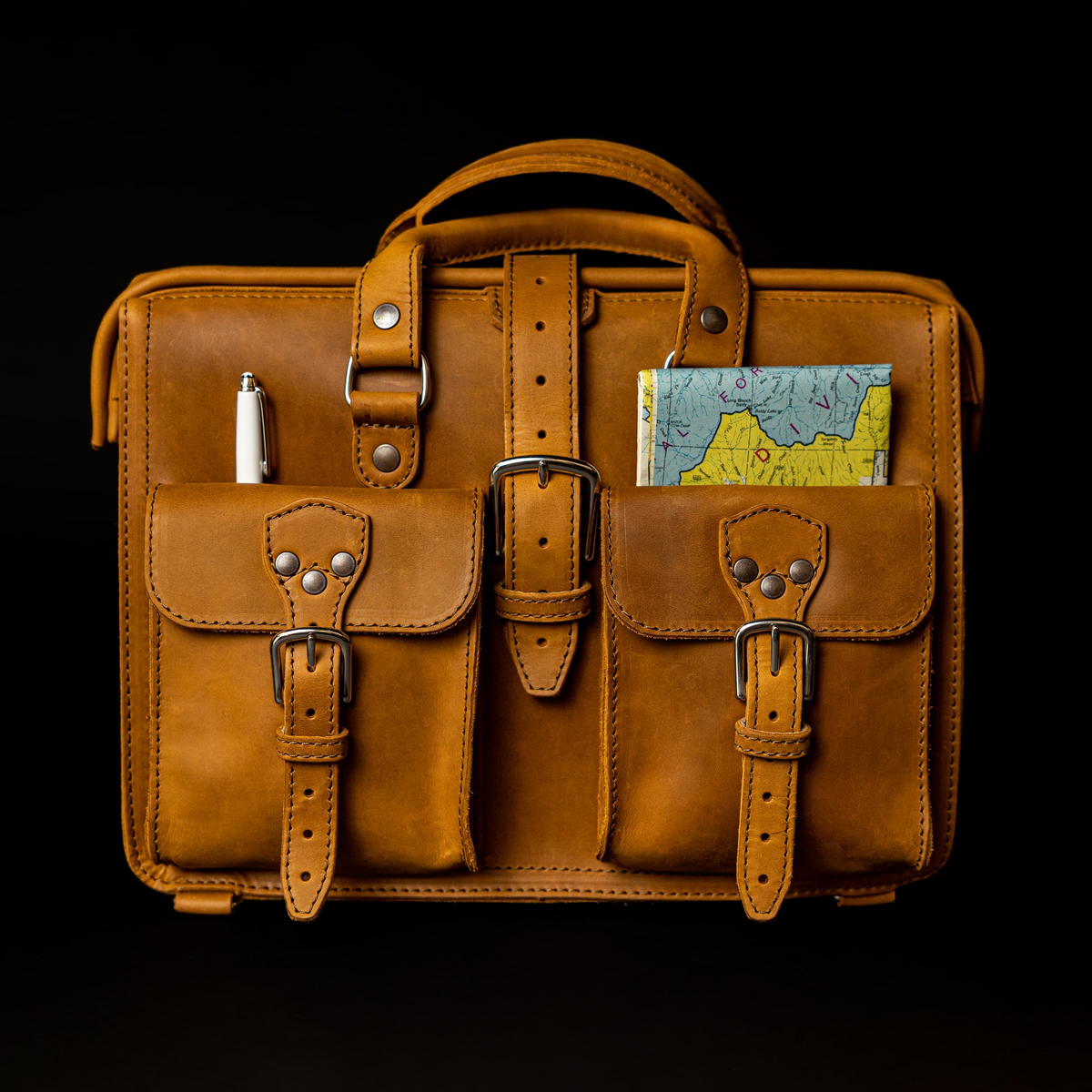 Saddleback Leather Flight Bag in Tobacco with front pockets holding a pen and a folded map, on a black background