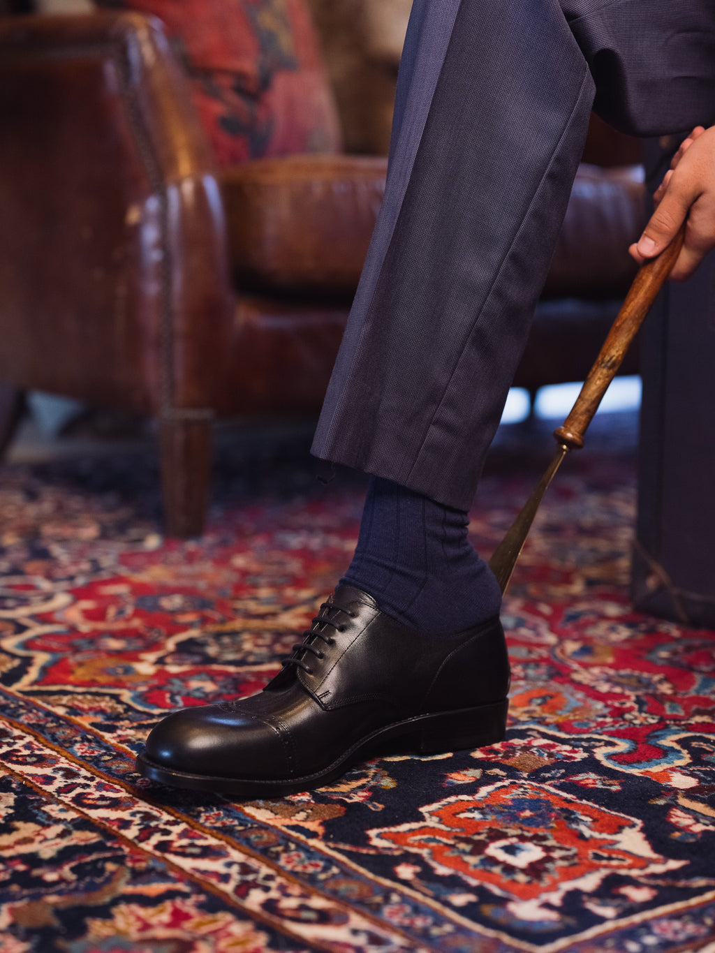 Toe Cap Shoes in Raven Black, single shoe put on with a wooden shoehorn beside a leather armchair