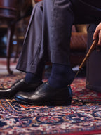 Toe Cap Shoes in Raven Black, worn with navy socks as a shoehorn eases the heel on a patterned rug