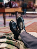 Toe Cap Shoes in Raven Black, worn with gray trousers, feet crossed and resting on a plaid blanket