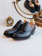 Toe Cap Shoes in Raven Black, front view of the pair on shoe trees beside a small brass clock