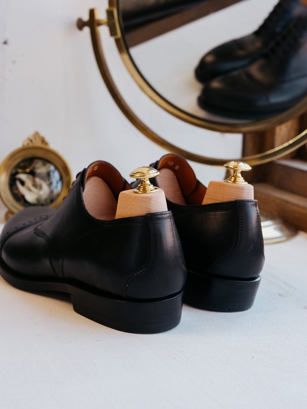 Toe Cap Shoes in Raven Black, rear view of the heels with shoe trees in front of a brass mirror