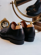 Toe Cap Shoes in Raven Black, rear view of the heels with shoe trees in front of a brass mirror