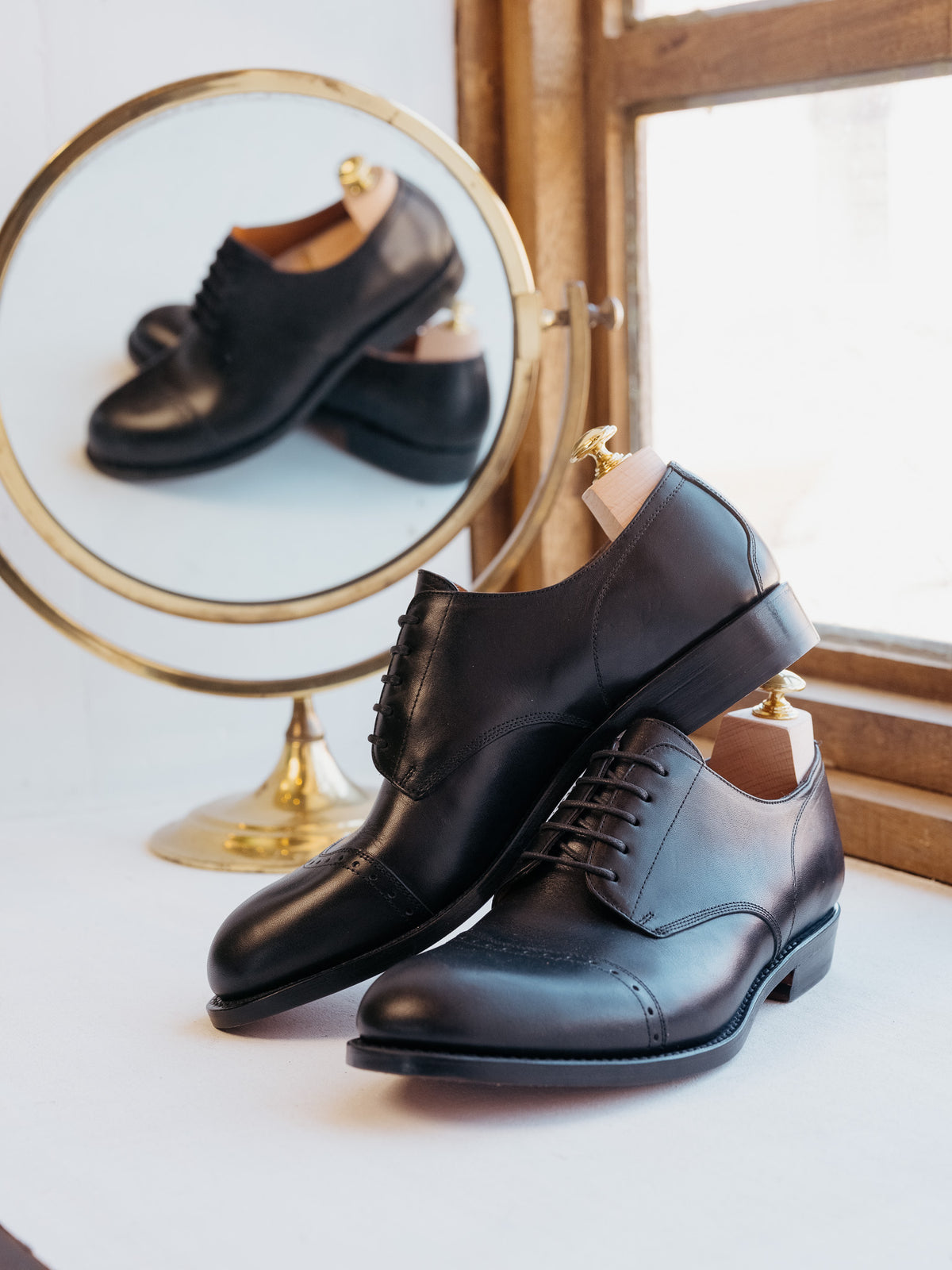 Toe Cap Shoes in Raven Black, pair on shoe trees beside a round brass mirror by a window