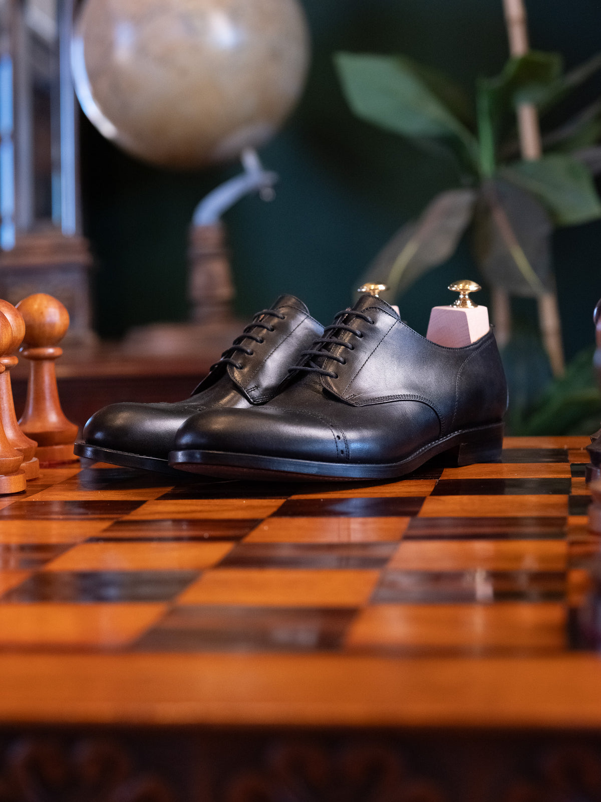 Toe Cap Shoes in Raven Black, pair with shoe trees on a chessboard before a vintage globe