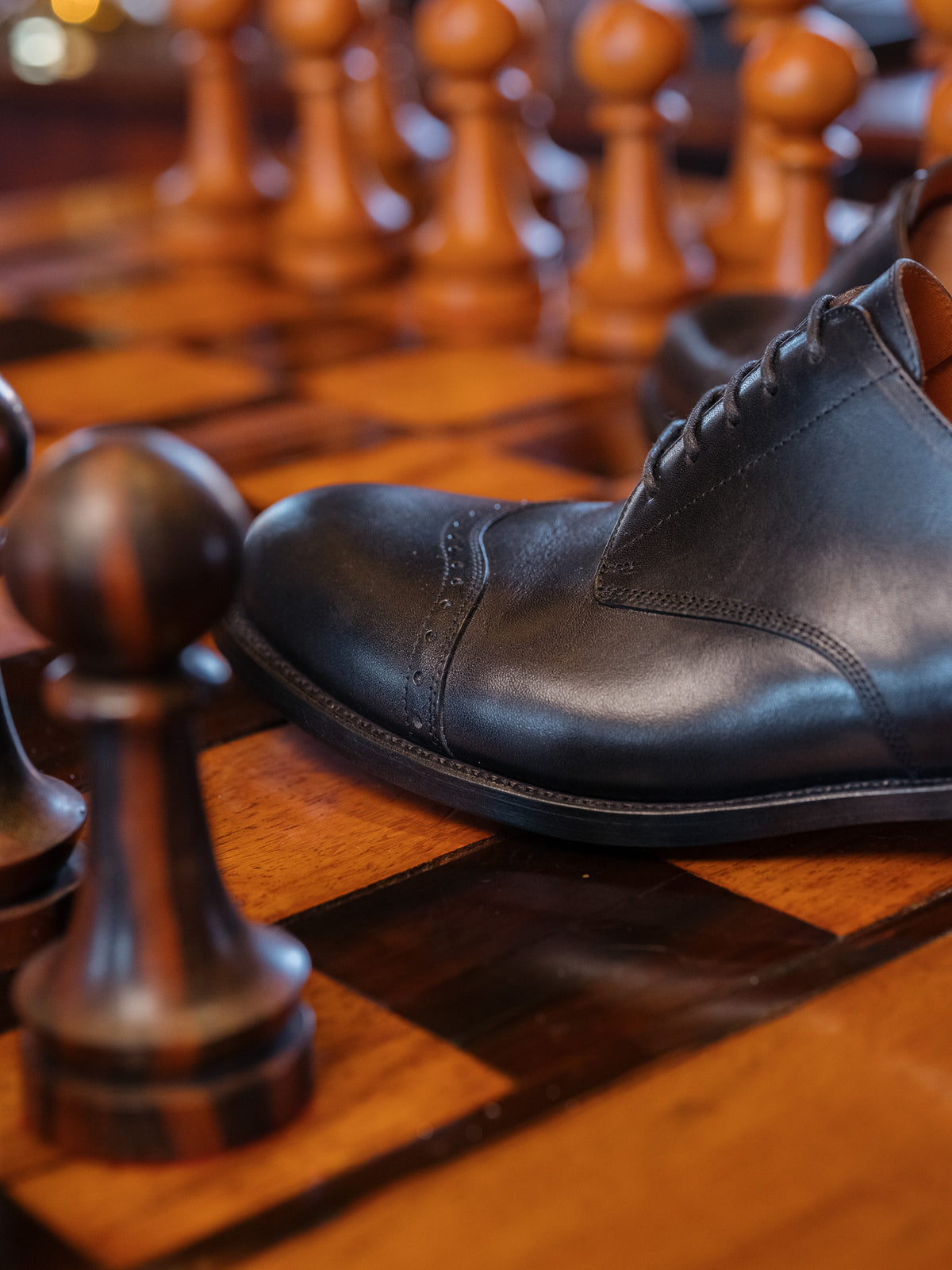 Toe Cap Shoes in Raven Black, close-up of a shoe resting on a wooden chessboard among chess pieces