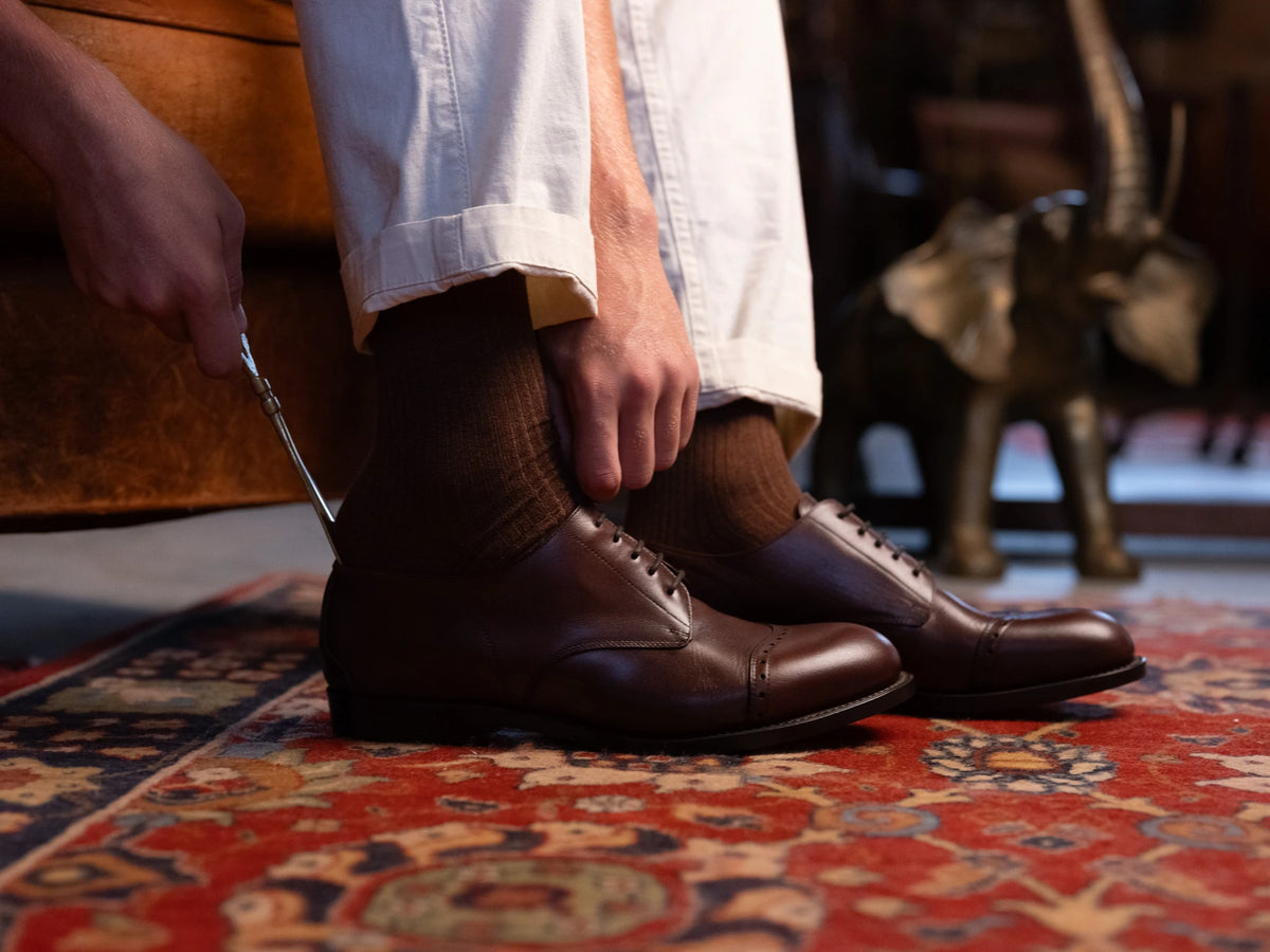 Toe Cap Shoes - Dark Oak, a person using a shoehorn to slip one on over a red rug beside an elephant statue