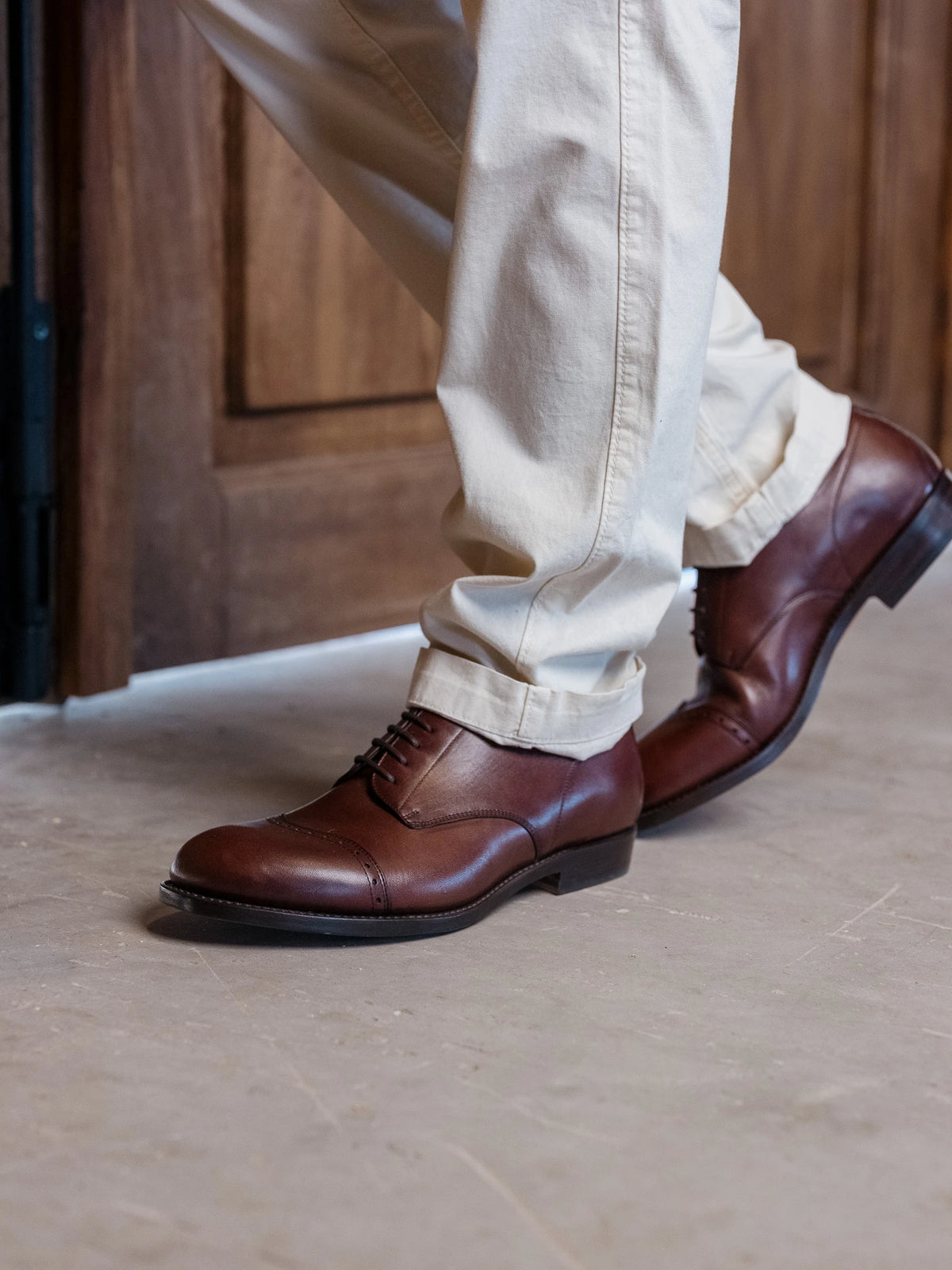 Toe Cap Shoes - Dark Oak on foot, side view of a step with cream trousers on a stone floor by a wooden door