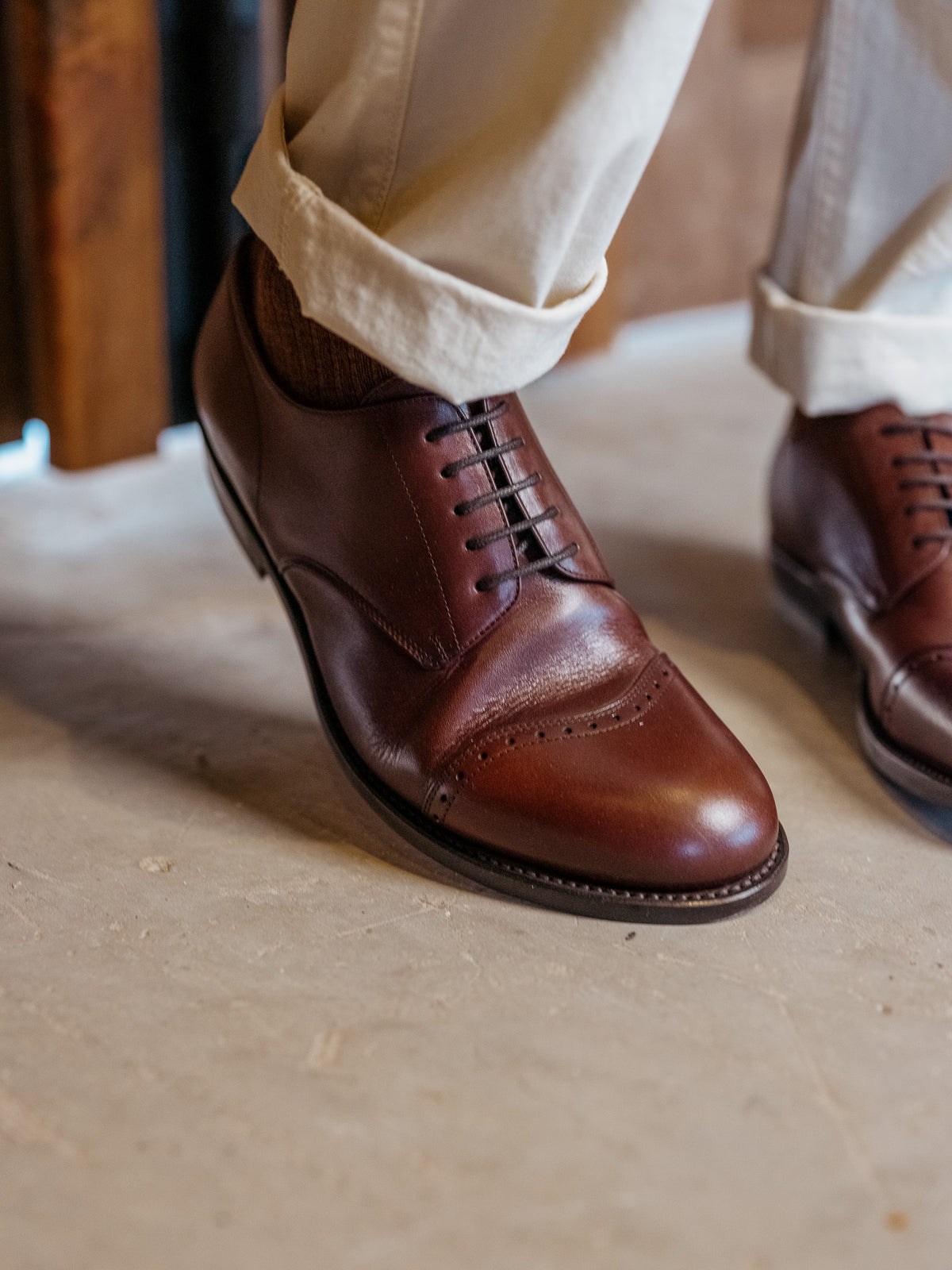 Toe Cap Shoes in Dark Oak, worn with cuffed cream trousers on a concrete floor