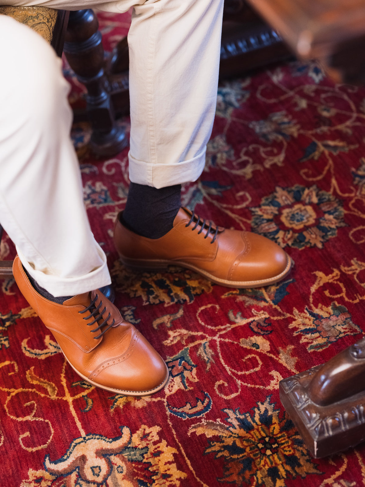 Toe Cap Shoes in English Tan, worn with cuffed cream trousers and dark socks on a red rug