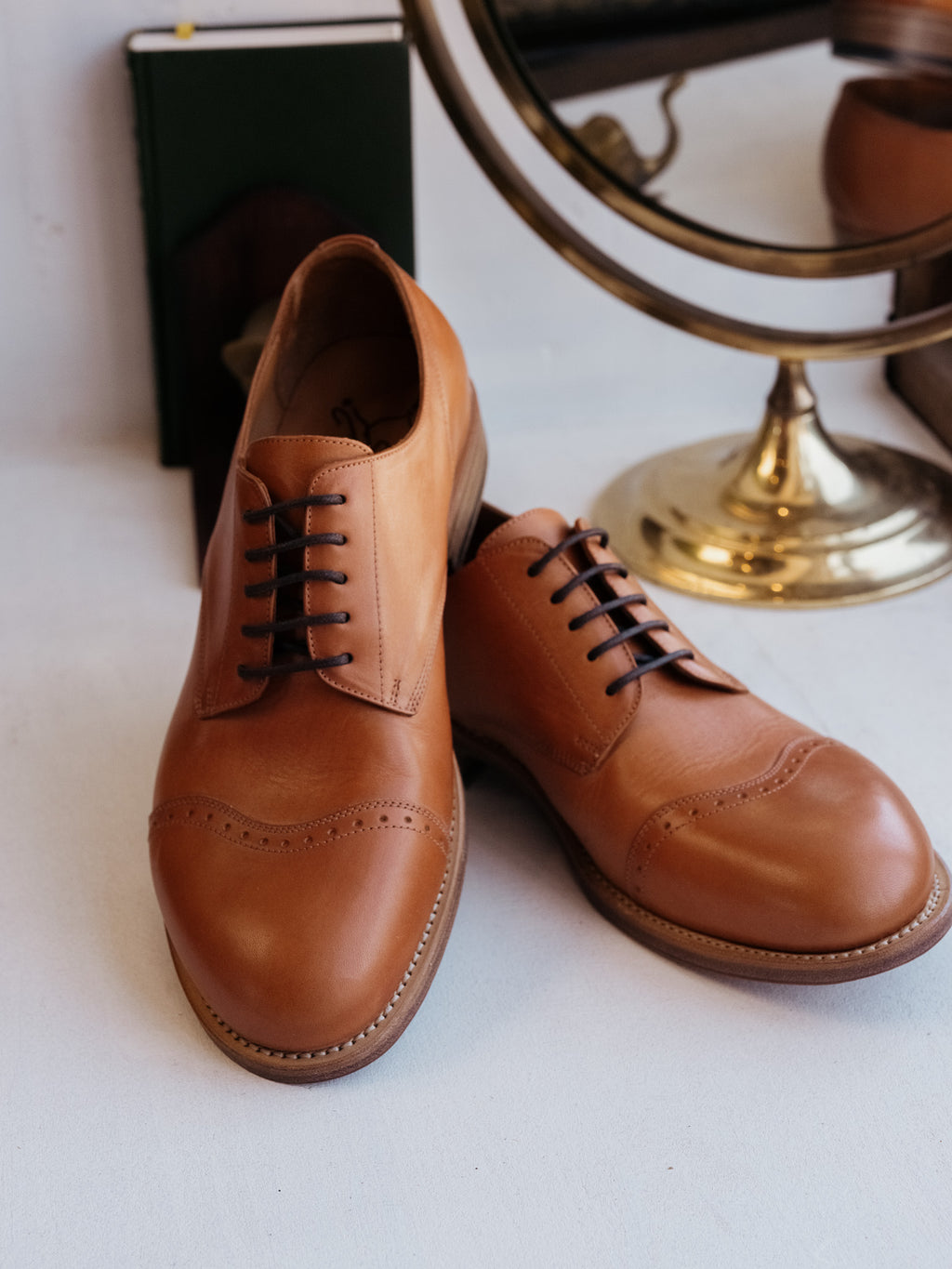 Toe Cap Shoes in English Tan, front three-quarter view of the pair beside a brass mirror and book