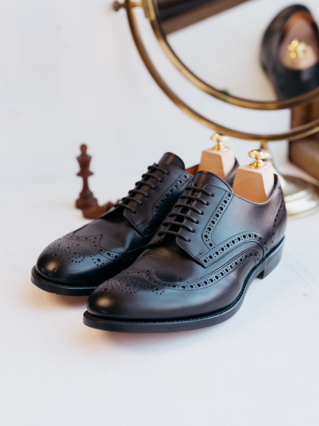 Wingtip Shoes in Raven Black, three-quarter view of the pair with shoe trees and brass mirror