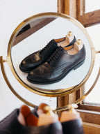 Wingtip Shoes in Raven Black, pair with shoe trees reflected in a round brass mirror