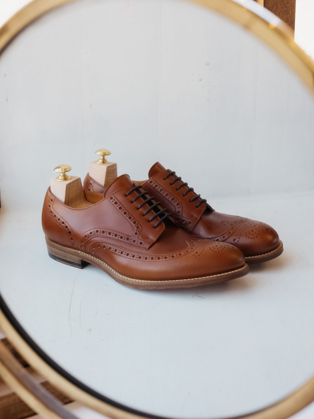 Wingtip Shoes in Chestnut, pair with shoe trees reflected in a round brass mirror