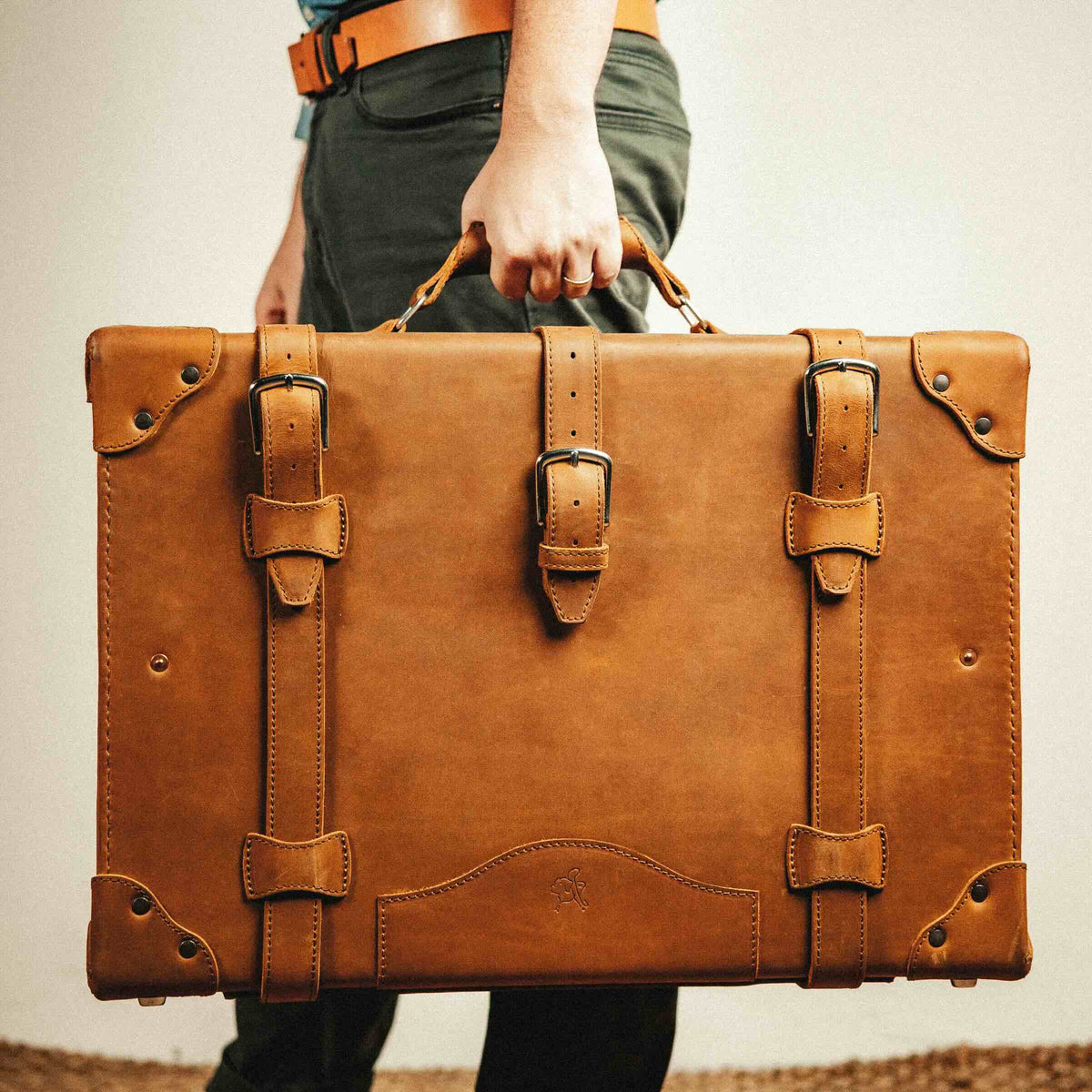 Grandfather Medium Suitcase in Tobacco carried by its top handle, showing scale against a man's leg