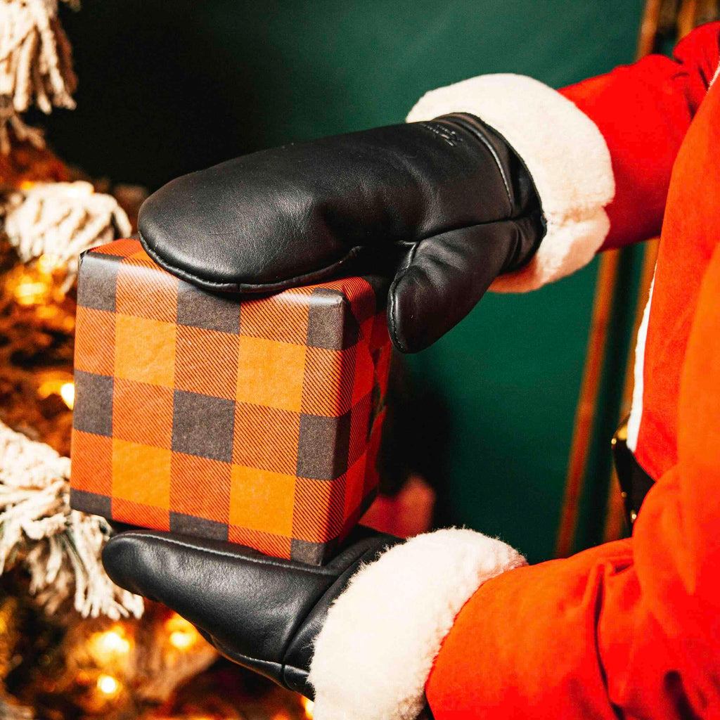 Sheepskin Mittens in black leather, worn by someone in a red Santa coat holding a plaid-wrapped gift