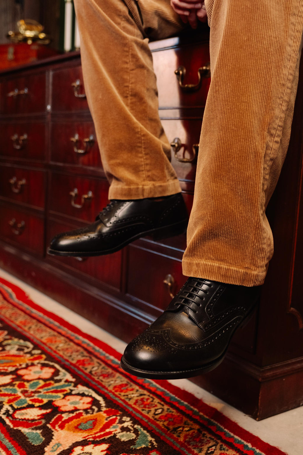 Wingtip Shoes in Raven Black, worn with tan corduroys while seated on a wooden chest