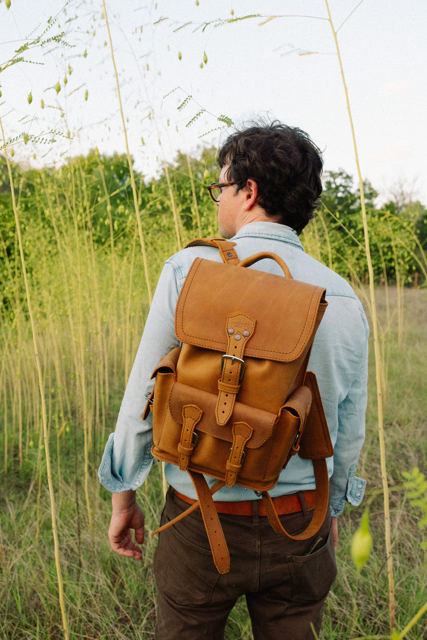 Small Front Pocket Backpack in Tobacco worn by a man in glasses looking over his shoulder among tall grass