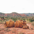 Little Big Mouth Toiletry Bag in Tobacco beside two larger leather travel bags on red dirt in a desert canyon