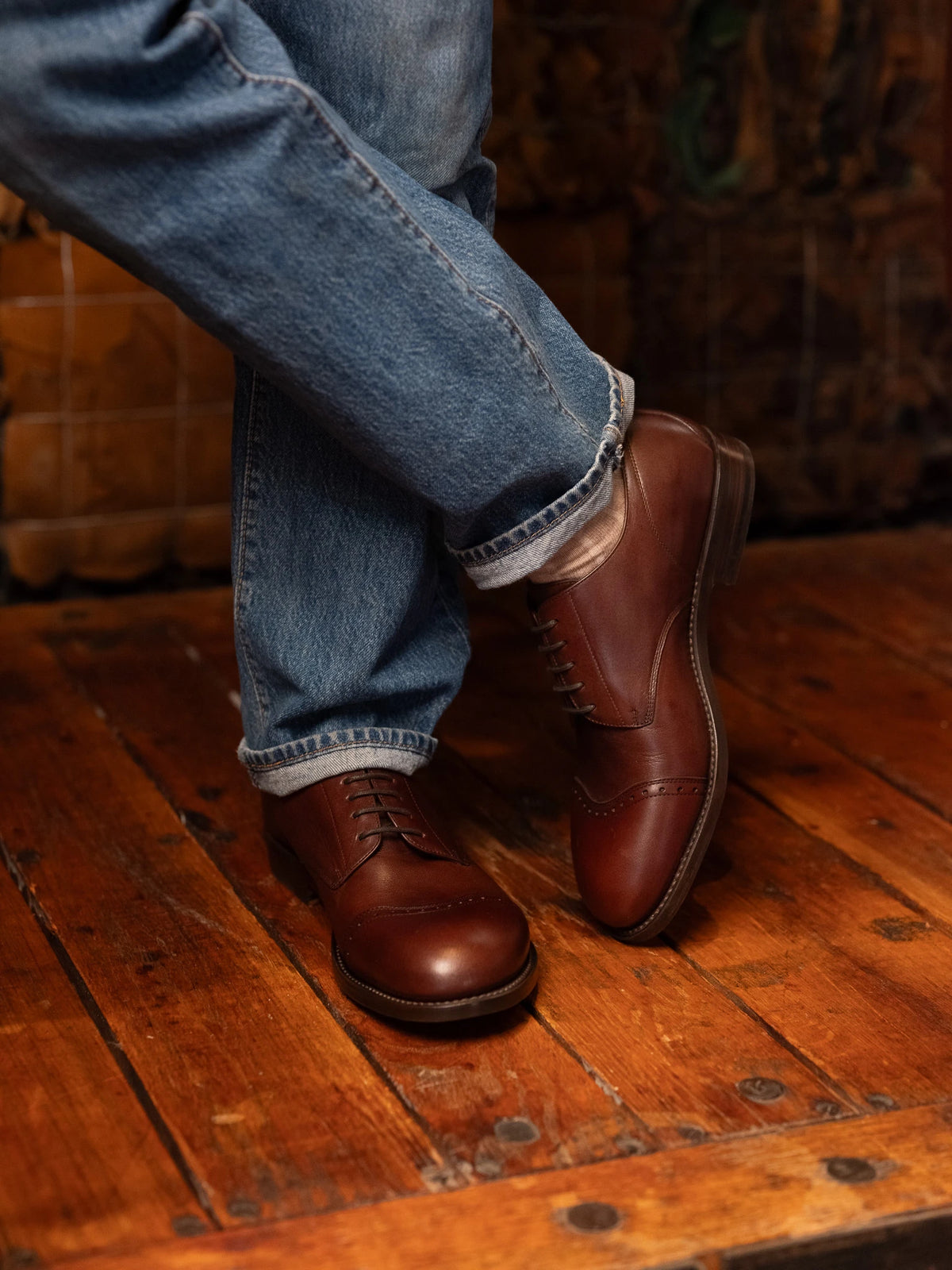 Toe Cap Shoes - Dark Oak worn with crossed legs and cuffed blue jeans on a rustic wood floor