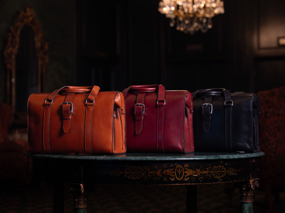 Three Silverback briefcases in tan, oxblood and black, lined up on an antique table in a dark panelled room beneath a lit chandelier.