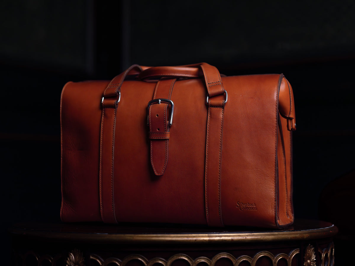 Spurgeon Briefcase in English Tan, front view with buckled strap on an ornate wooden pedestal in low light