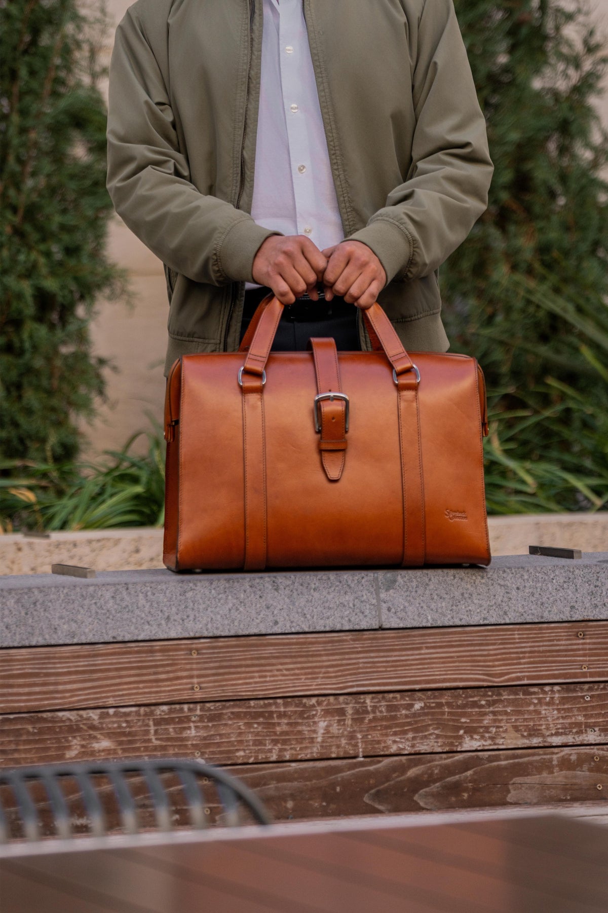 Spurgeon Briefcase in English Tan held by its top handles in front of a man in an olive jacket outdoors