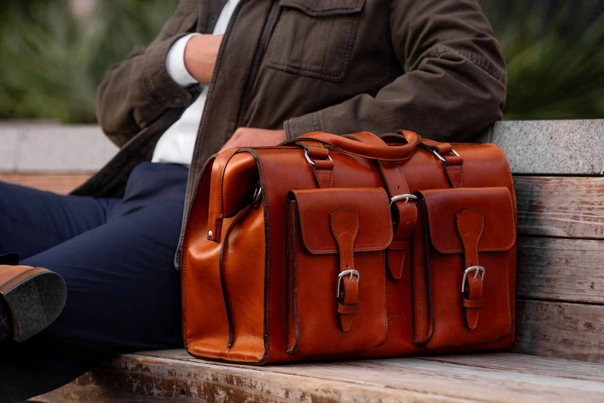 A tan leather briefcase with buckled front pockets resting on a wooden bench beside a seated man in a dark jacket.