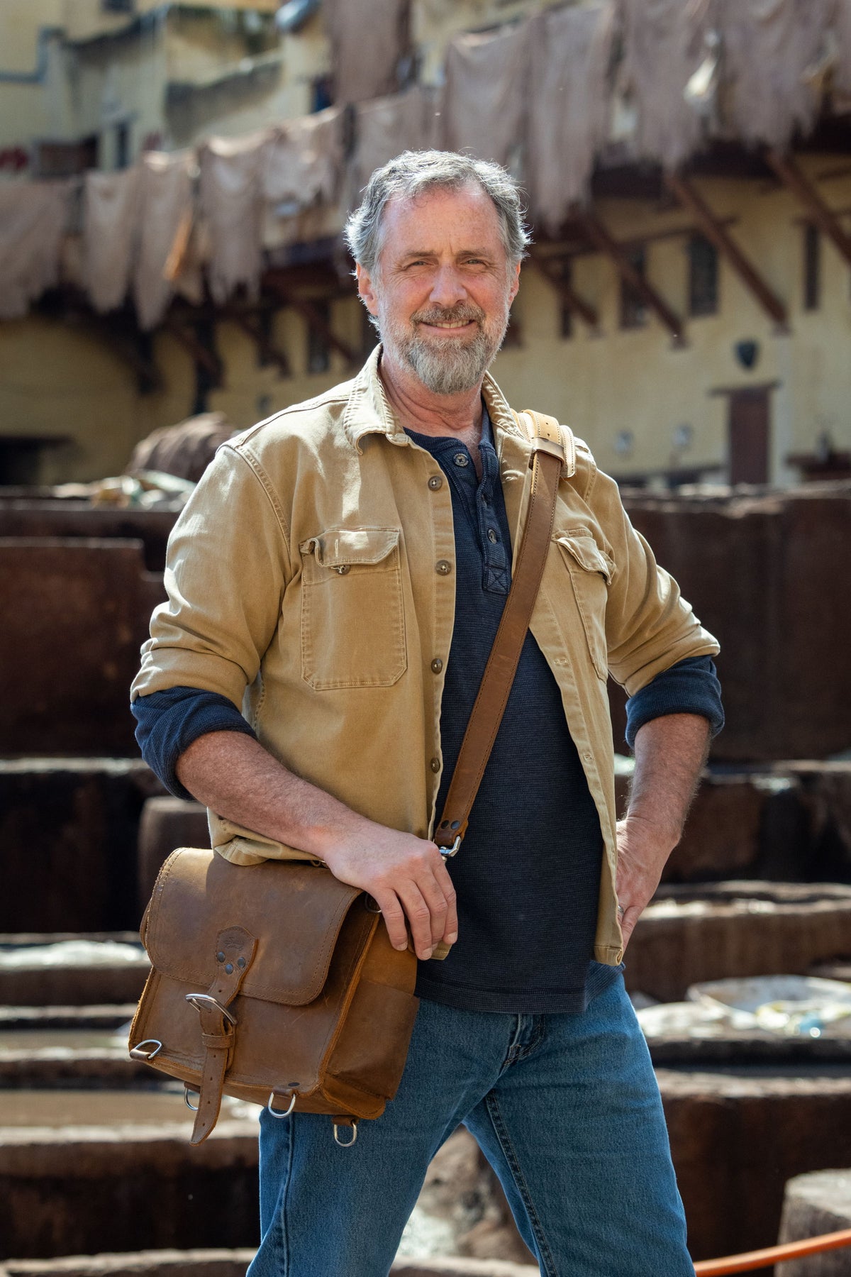A bearded man in a tan work jacket standing at a leather tannery, with stone dye pits and hides drying behind him, a worn brown satchel on his shoulder.
