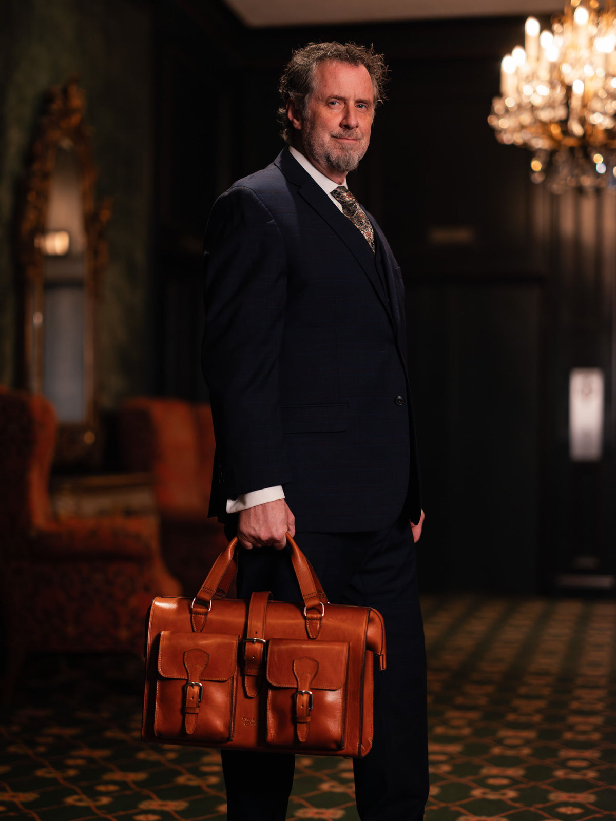 A man in a dark three-piece suit carrying a tan leather briefcase through a chandelier-lit hotel lounge.