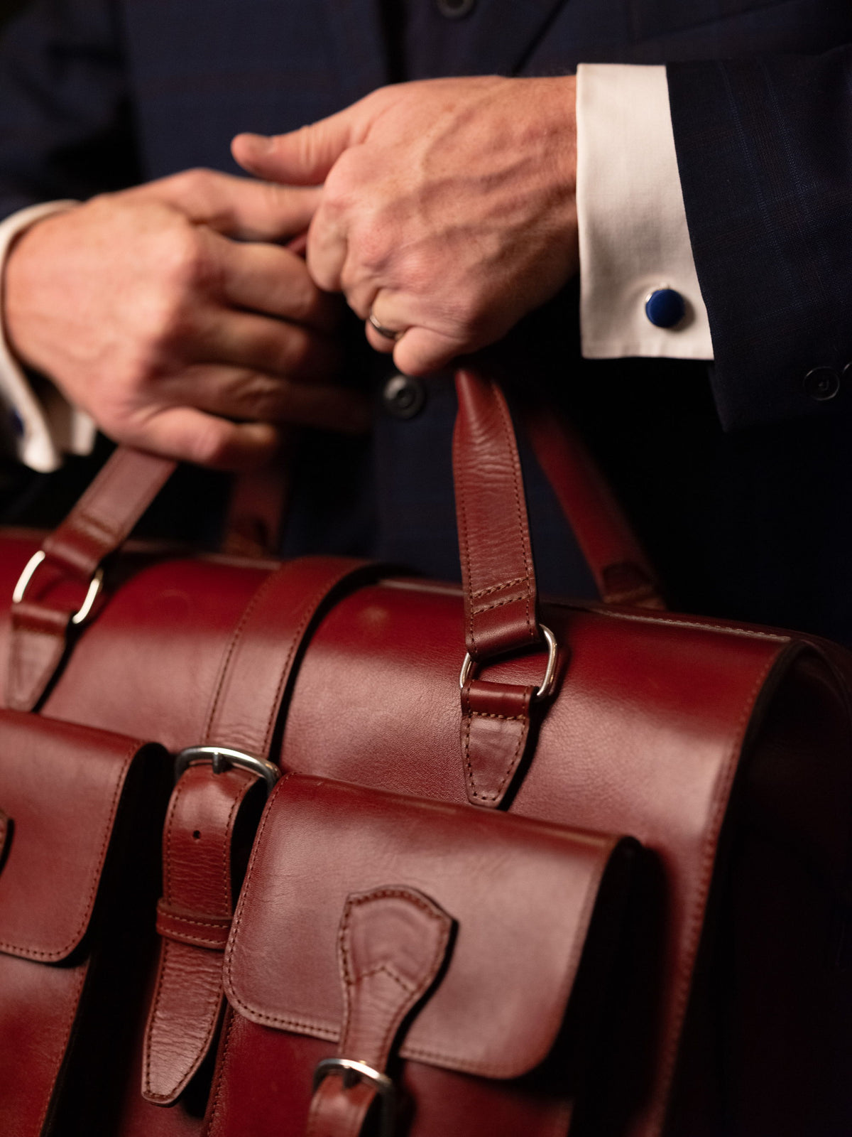 Close-up of a man's hands, one shirt cuff fastened with a blue cufflink, gripping the handles of an oxblood leather briefcase.