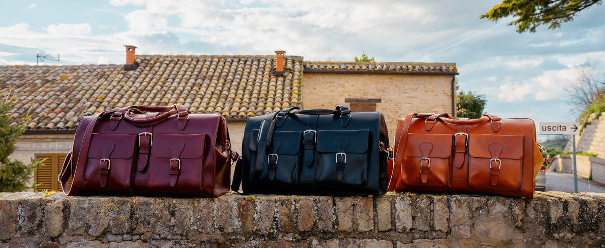 Wesley Deep Pocket Duffle in Oxblood, Raven Black and English Tan side by side on a stone wall under a cloudy sky