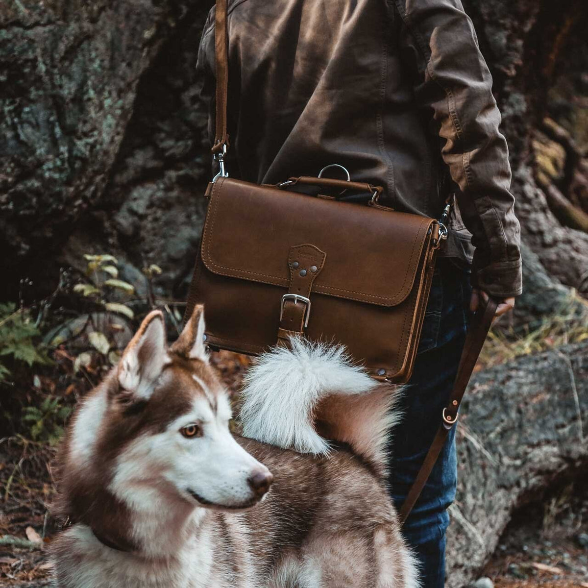 Hiker carrying a brown leather slim briefcase on a shoulder strap beside a husky on a rocky trail