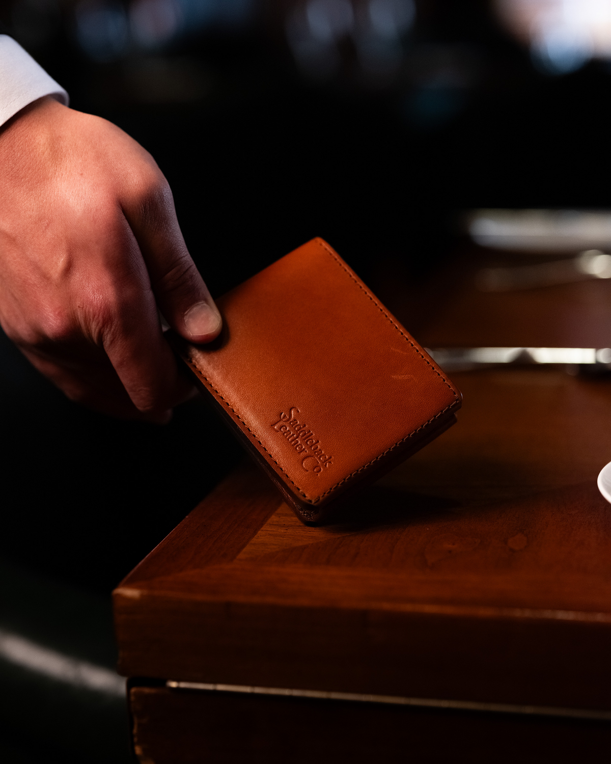 Hand placing a natural brown Silverback leather bifold wallet on the edge of a dark wooden table