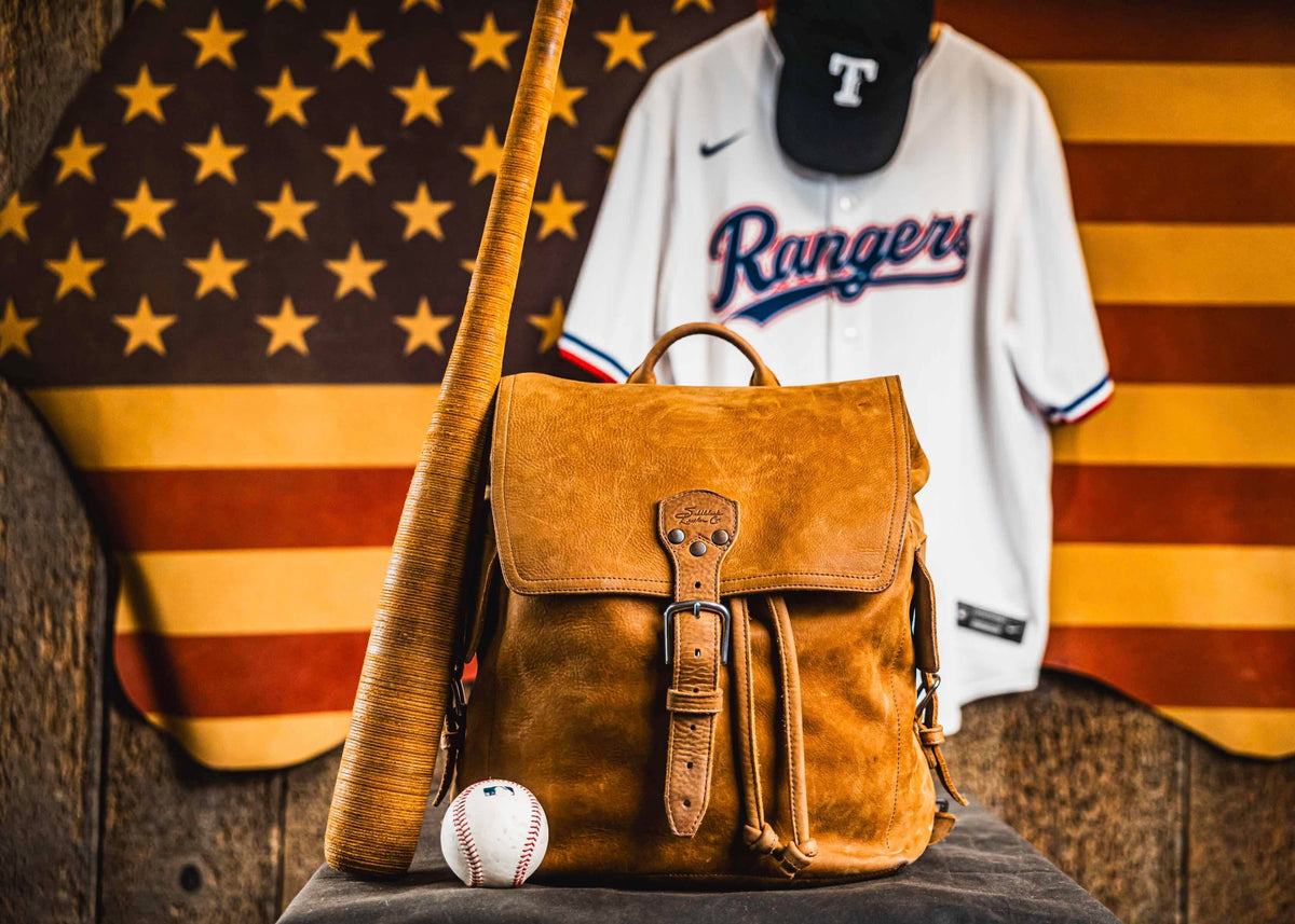 A tan leather backpack with a bat and a baseball, in front of a Rangers jersey and cap hung on an American flag.