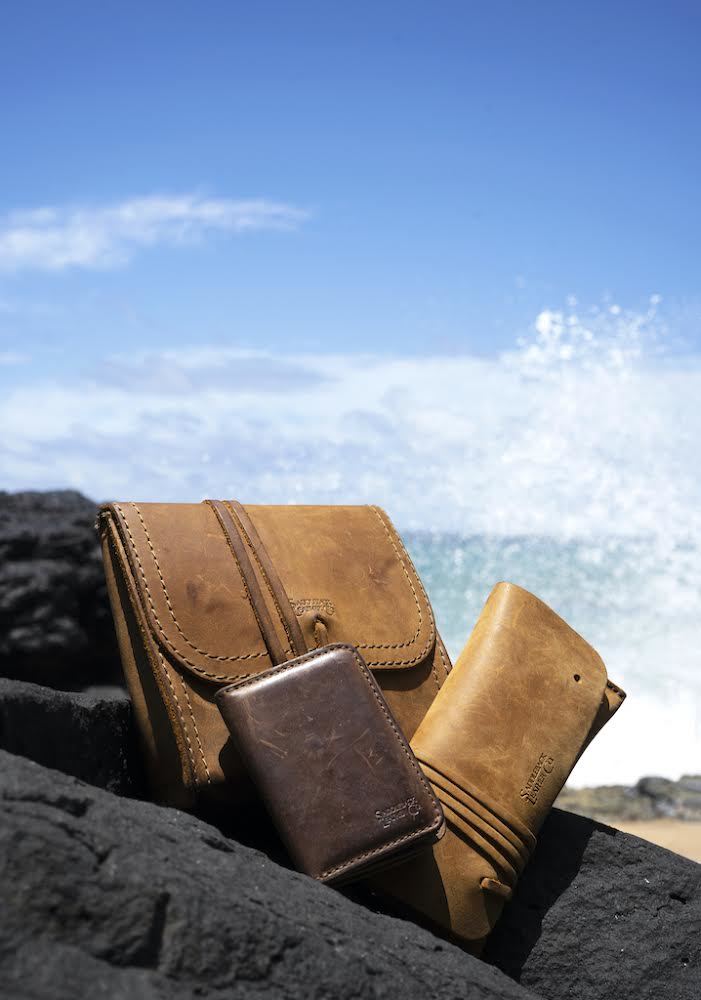 Cash & Receipt Bag in Tobacco on black lava rocks beside other leather goods, ocean waves splashing behind
