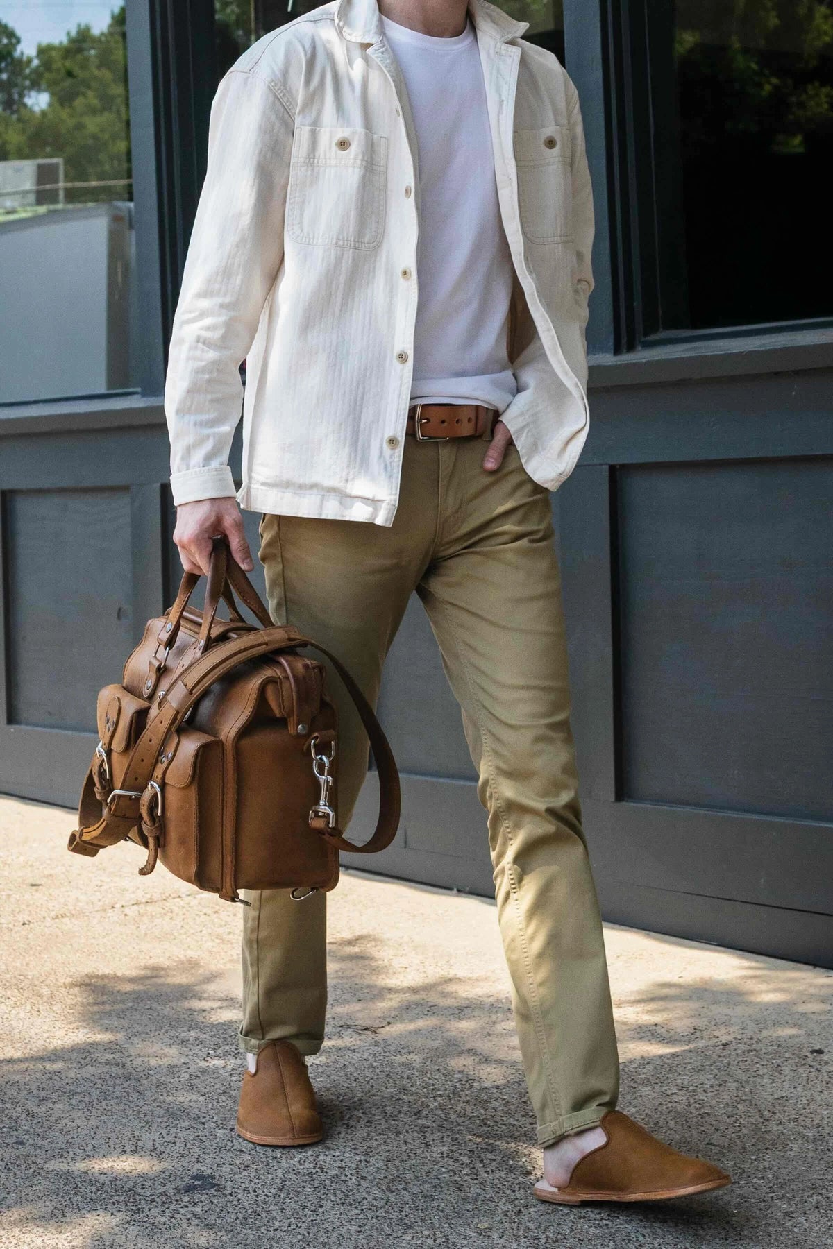 Man in a cream jacket and khakis wearing tan leather barefoot mules, carrying a brown leather duffle bag
